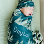 Newborn baby wrapped in a teal swaddle with trees and 'Dayton' printed on it, wearing a matching hat.