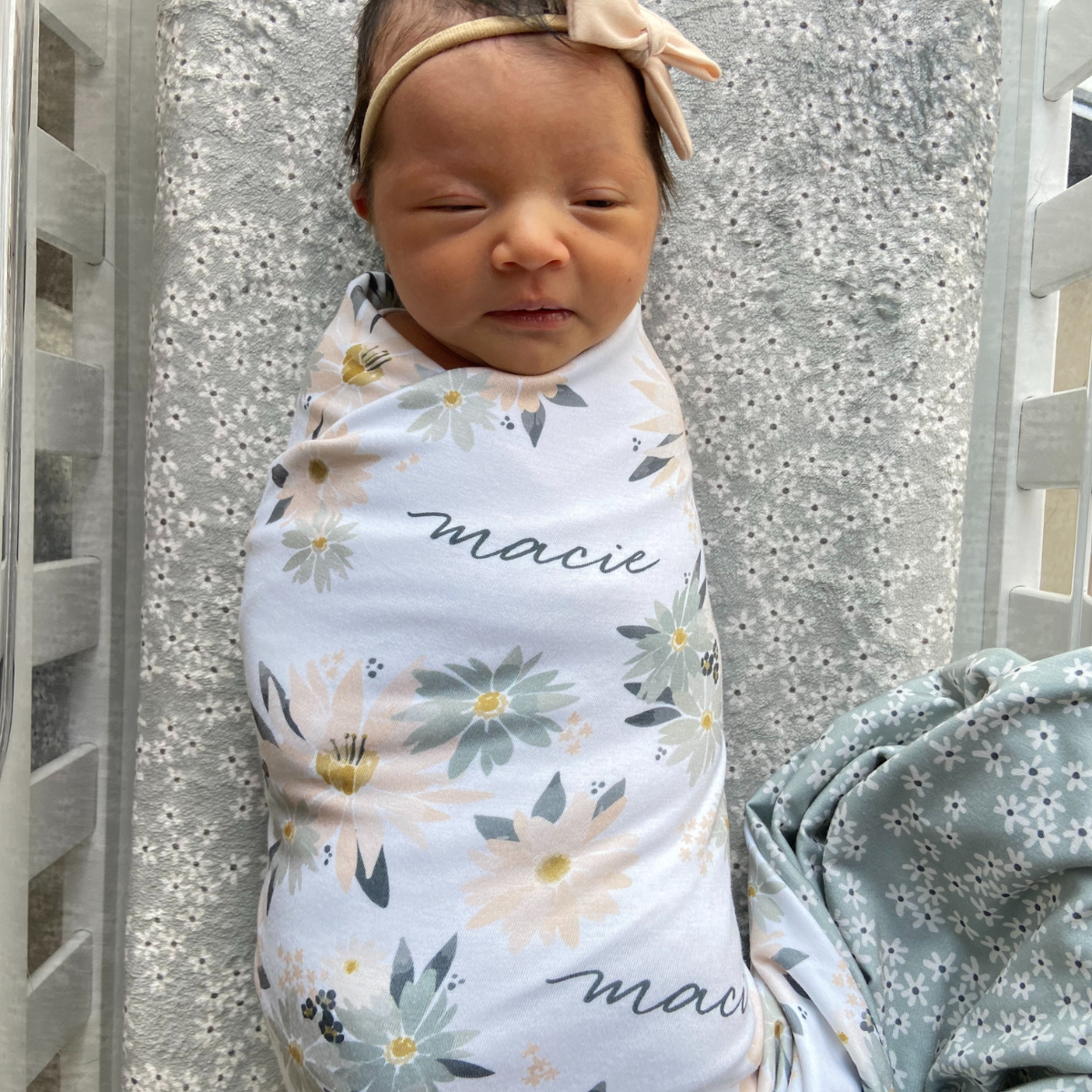 Newborn baby wrapped in a floral swaddle with name 'macie' printed on it, laying on a crib with floral crib sheet