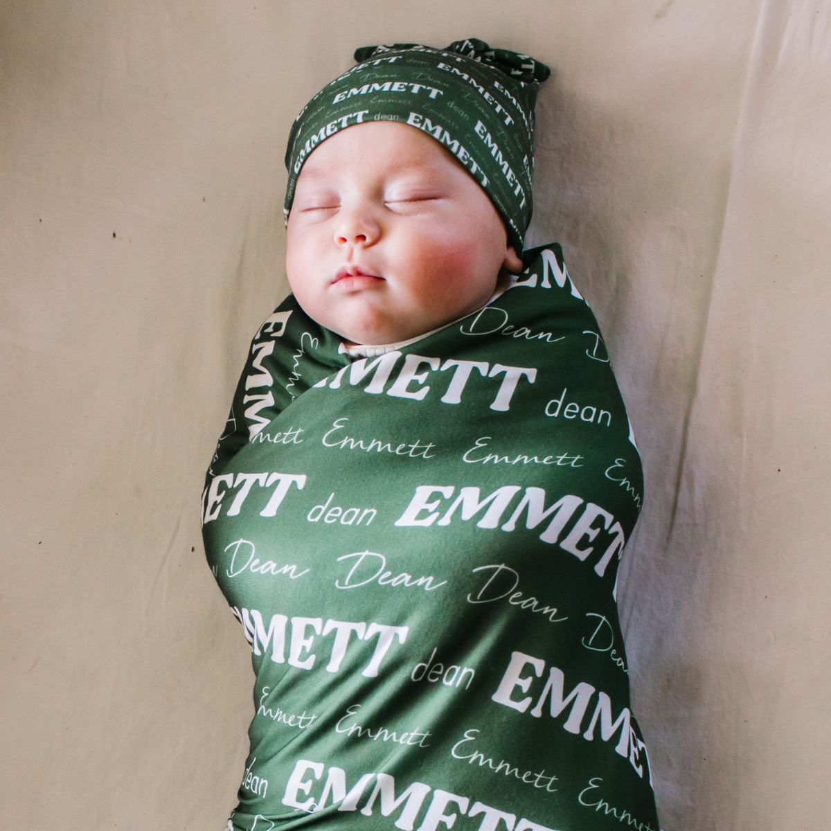 Newborn baby wearing personalized baby swaddle with name on it in white font on a dark green swaddle.
