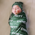 Newborn baby wearing personalized baby swaddle with name on it in white font on a dark green swaddle.