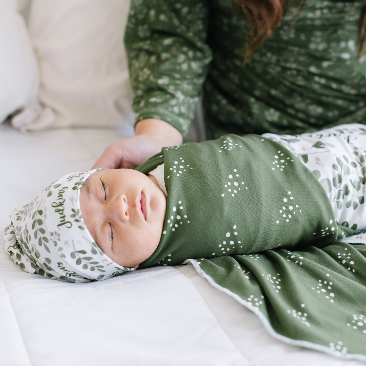 Baby wrapped in a green and white personalized name swaddle with floral patterns on a white background