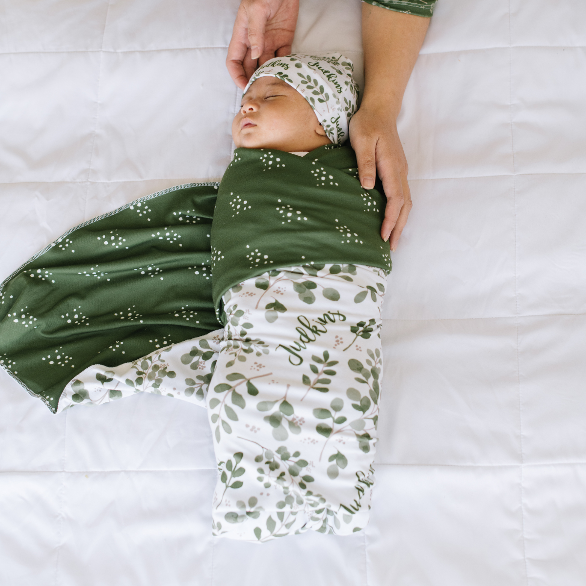 Newborn baby wrapped in a green personalized name swaddle with floral patterns on a white background