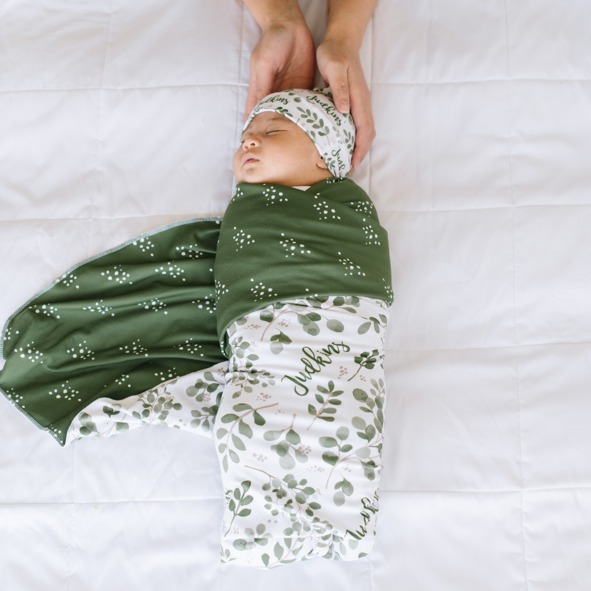 Baby wrapped in a green and white personalized name swaddle with floral patterns on a white background